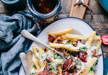 bacon strips and melted cheese topped fries on oval white and blue platter with gray stainless steel forks